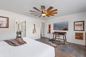 Un dormitorio con una cama grande, un ventilador de techo, una guitarra, un escritorio con una silla y un cuadro en la pared.