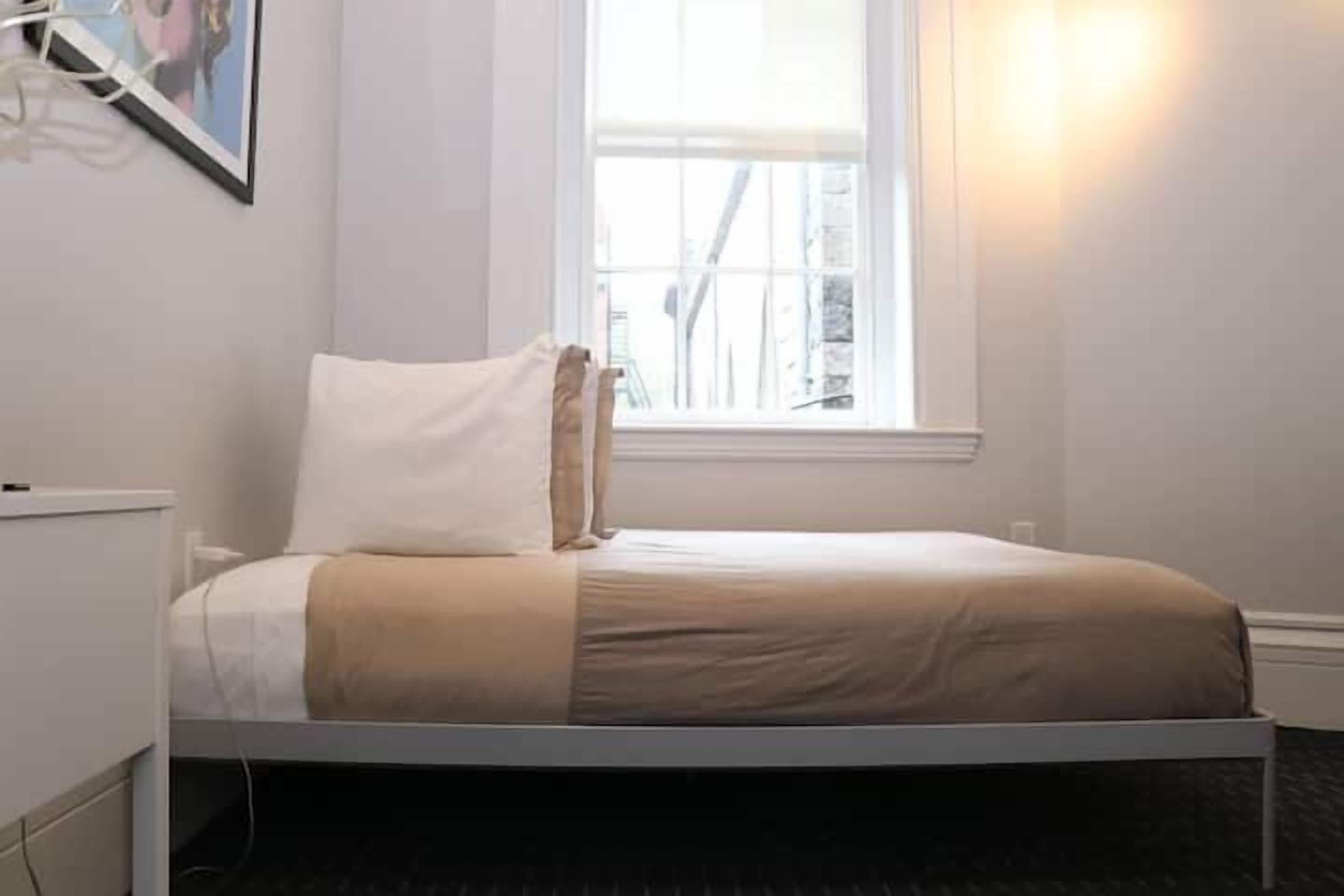 Ein Einzelbett mit weißem Kopfteil, einer beigen Decke und einem Kopfkissen, platziert unter einem Fenster mit Blick auf Gebäude.