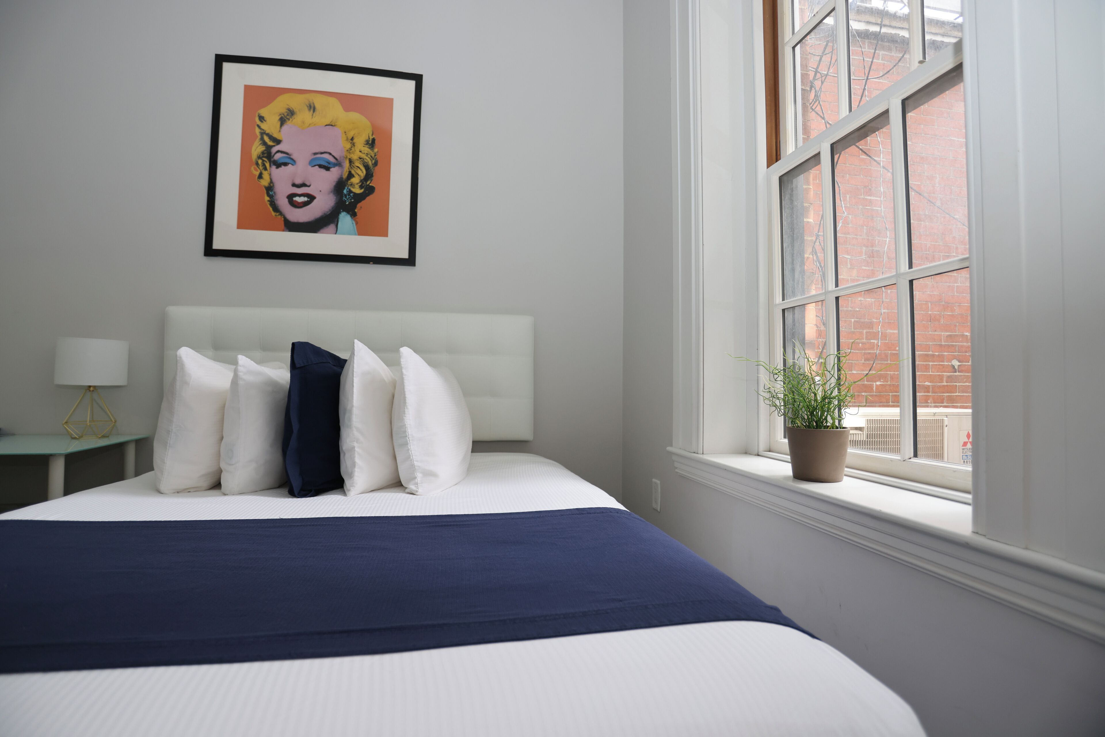 Un letto rifatto con cura, con copriletto blu, cuscini bianchi e un quadro incorniciato sopra.