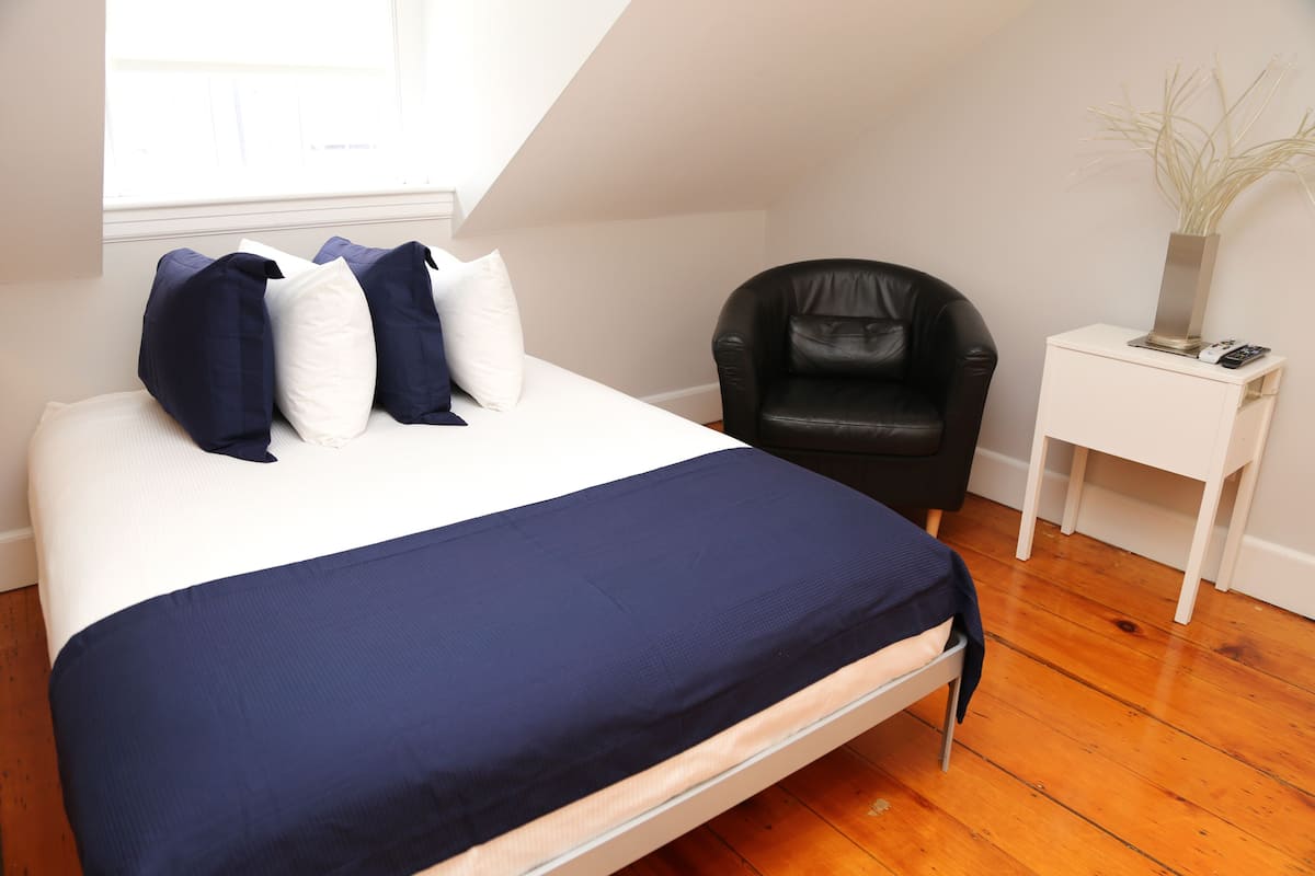A bedroom with a bed, a black leather chair, a white bedside table with a vase, and a window with blinds.