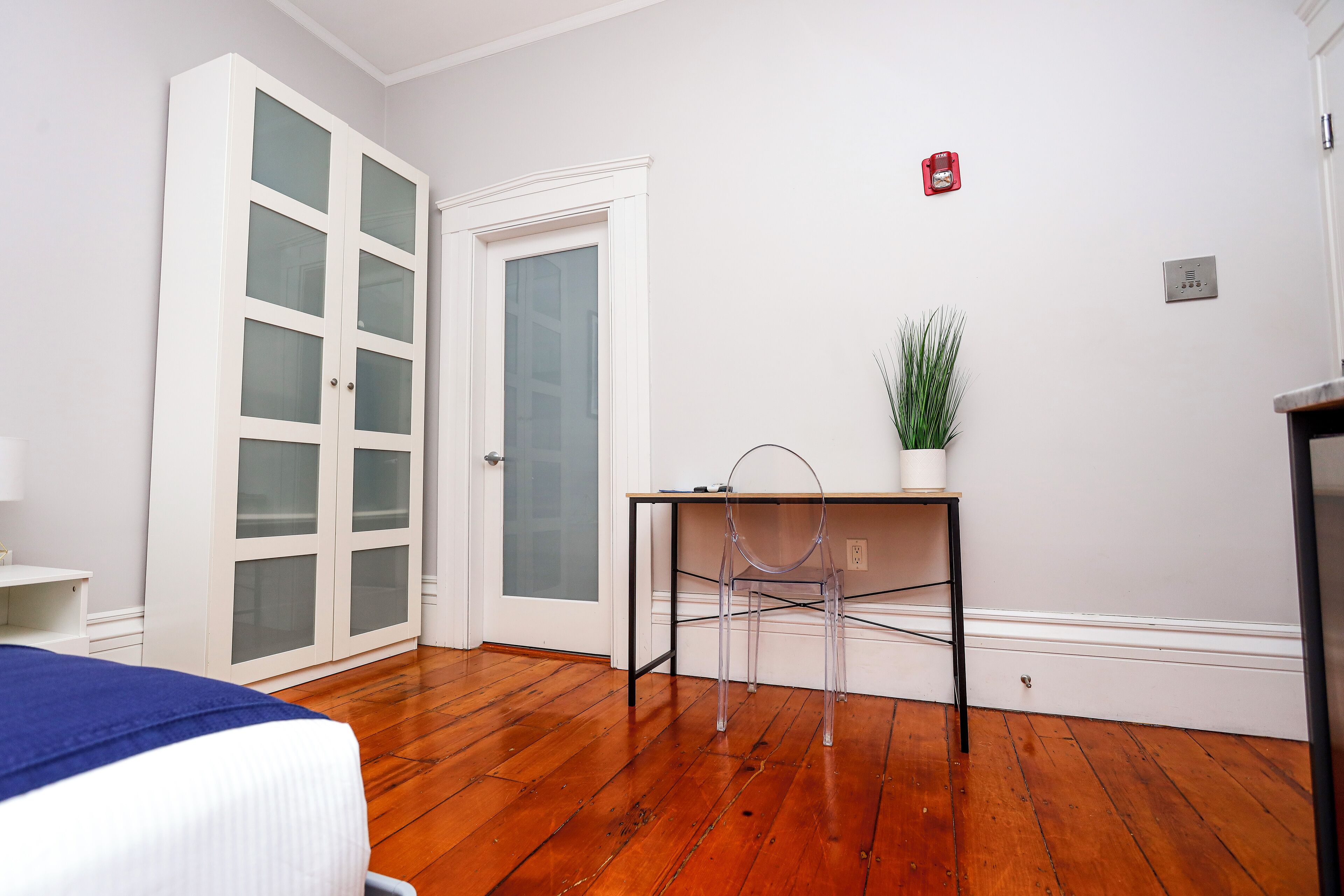 Une chambre comprenant un lit, un bureau avec une chaise, une plante en pot et une armoire blanche avec des portes en verre.