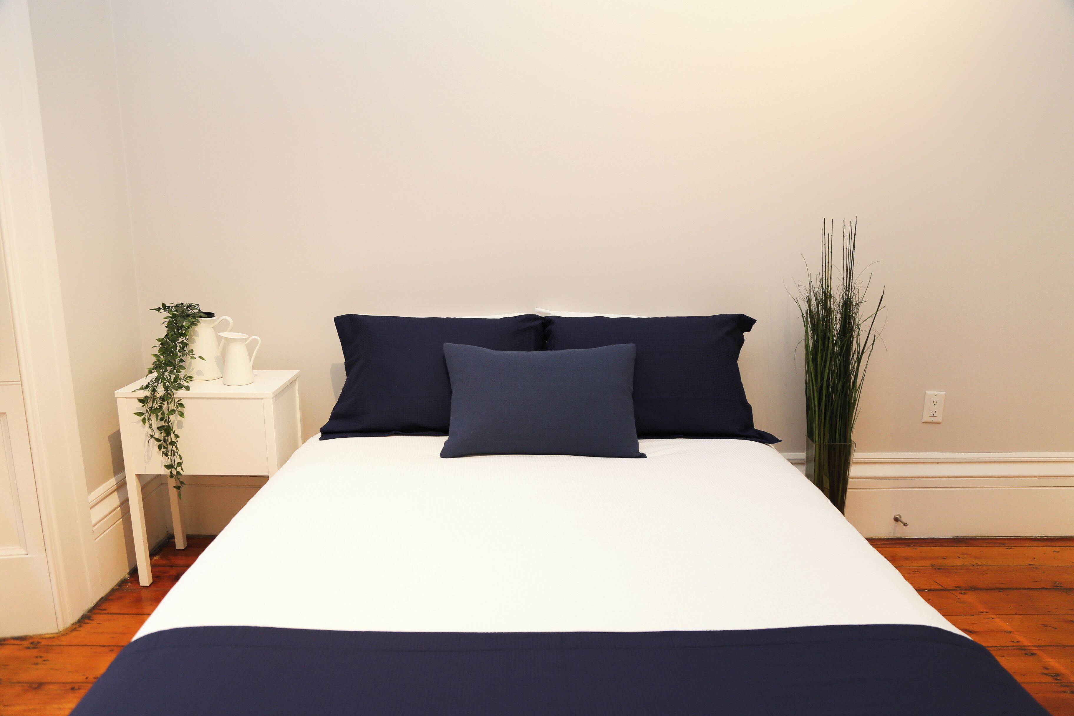 A neatly made bed with white and navy bedding, a small bedside table with a plant, and a tall vase of greenery.