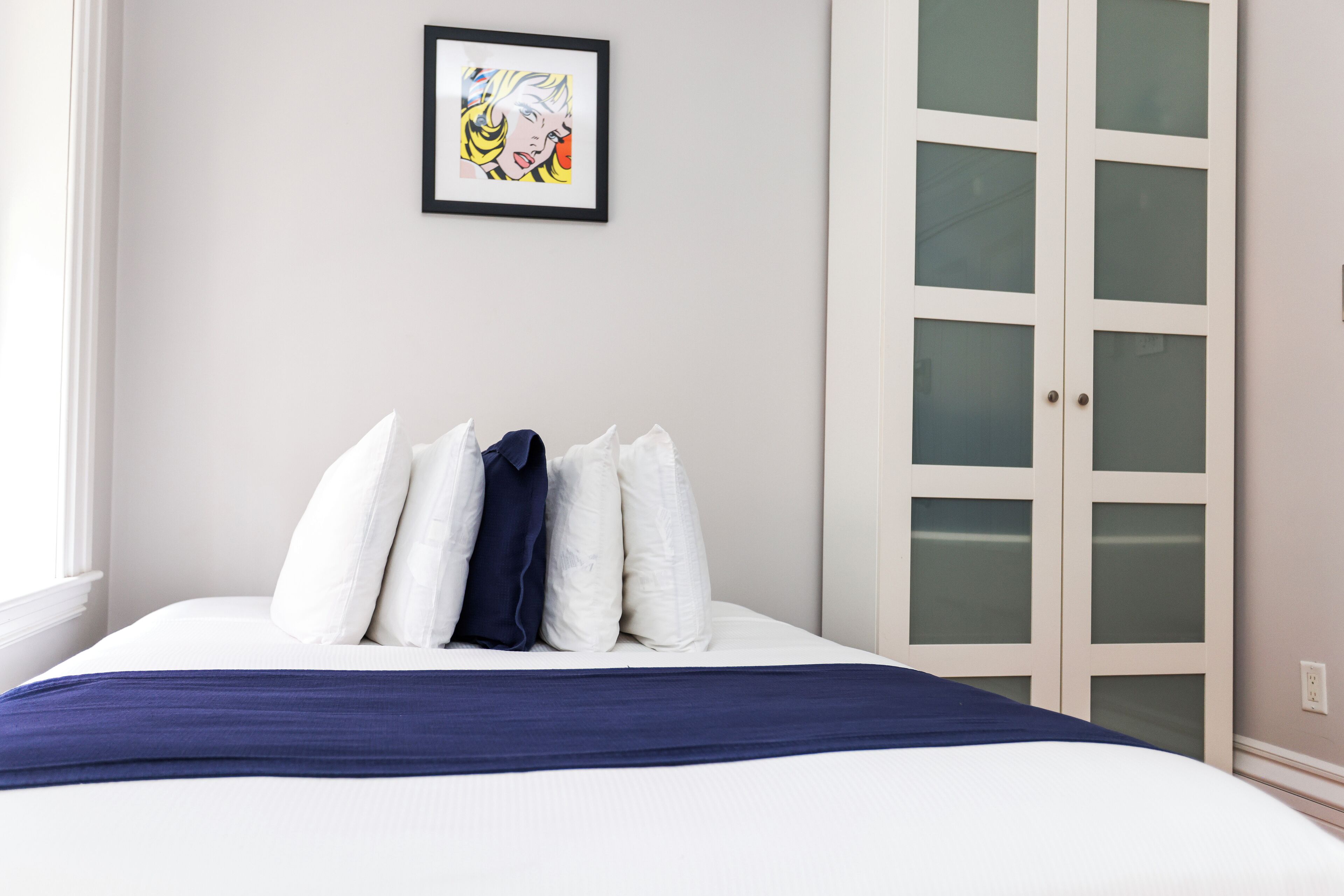 Una cama bien hecha con cubrecama azul y almohadas blancas, un cuadro enmarcado en la pared y un armario con puerta de vidrio.