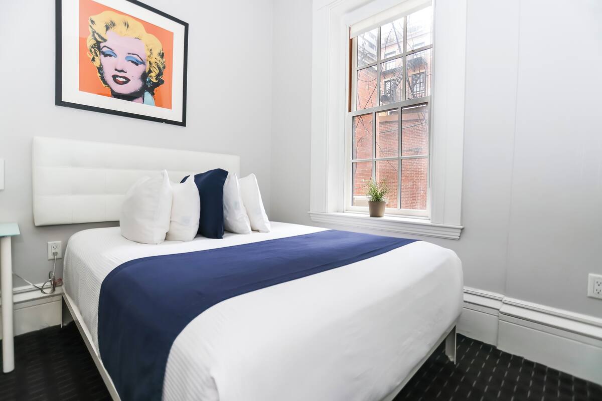 A neatly made bed with white and blue bedding, a framed artwork above it, and a window with a view of a brick building.