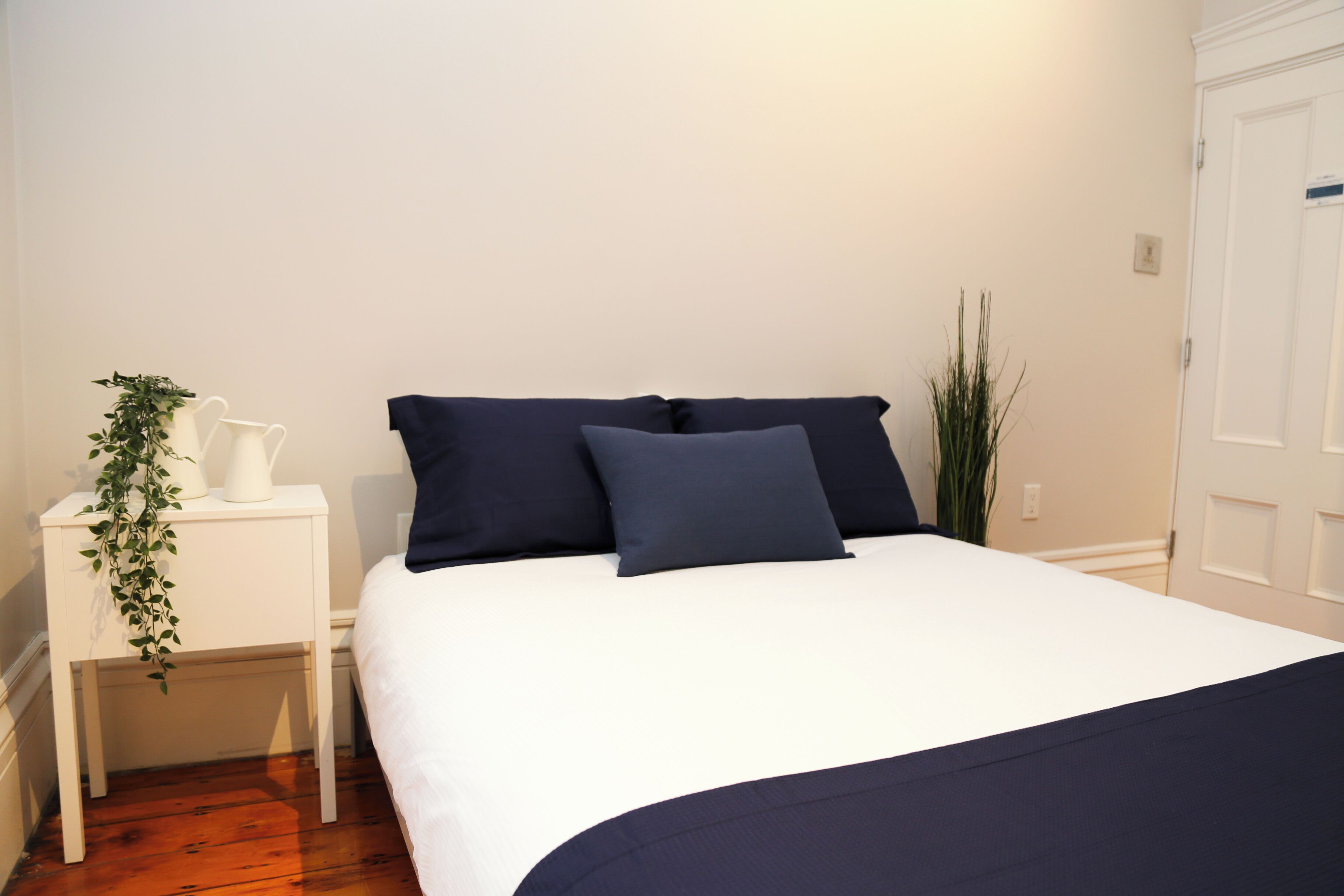 A neatly made bed with white and navy bedding, a white nightstand with a plant, and a door to the right.