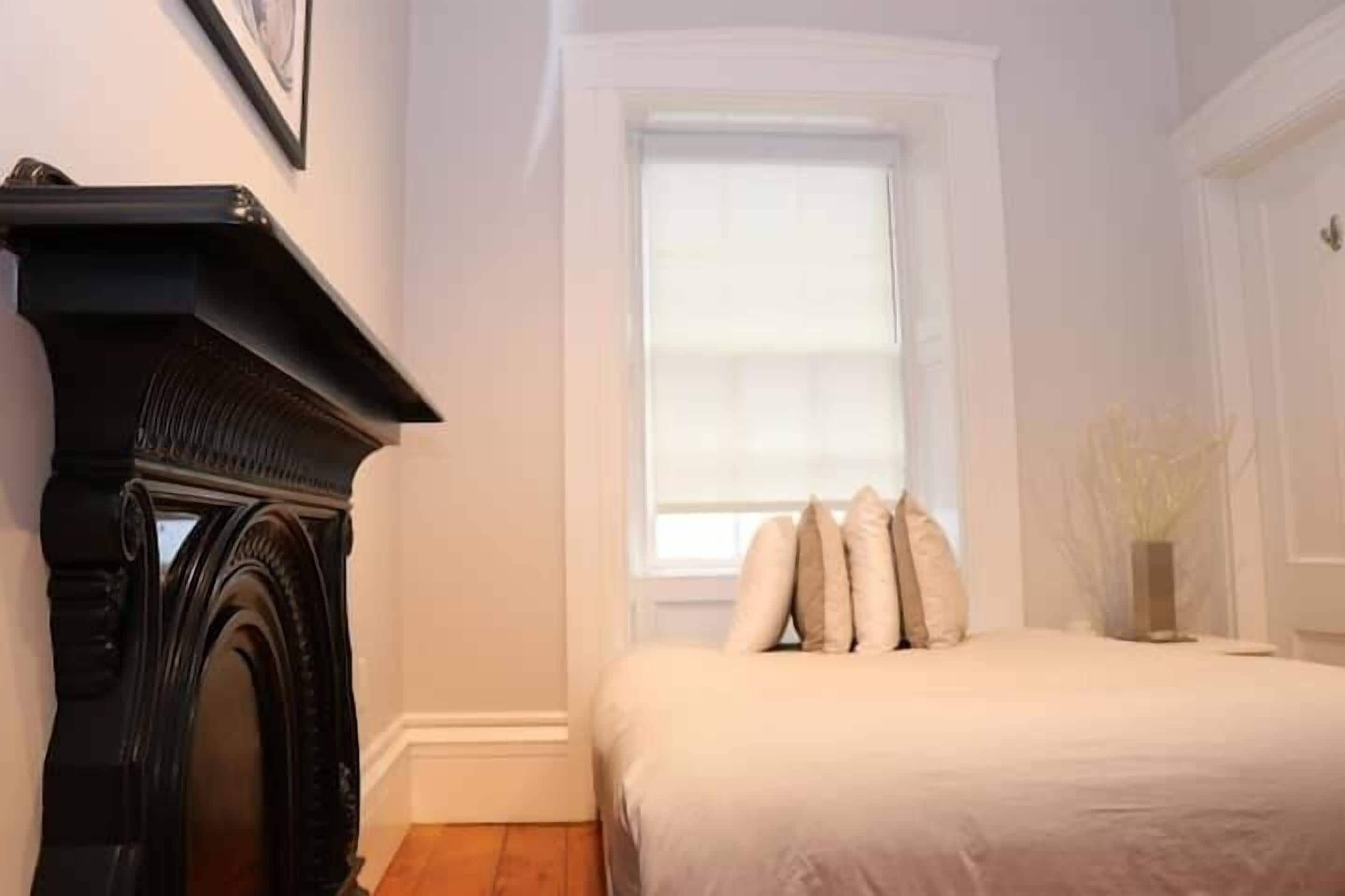 A bedroom with a fireplace, a window with blinds, a bed with pillows, and a vase with dried plants.