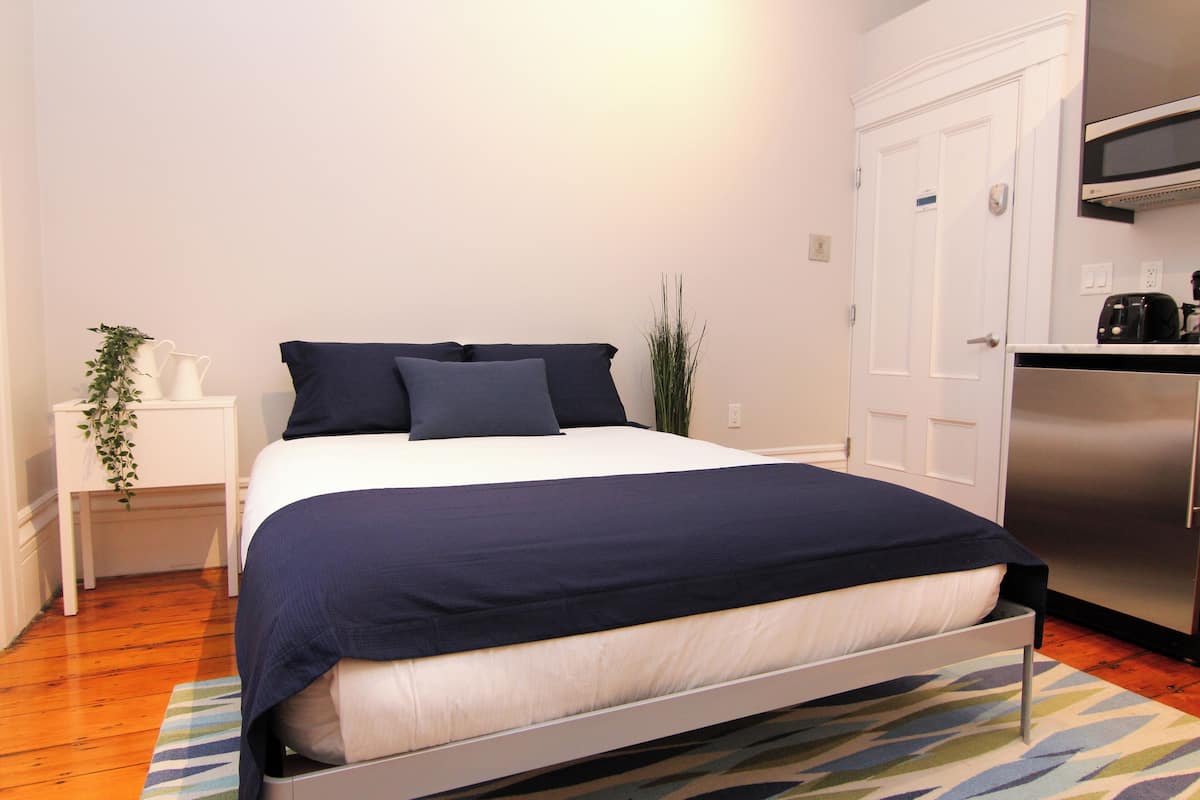 A bedroom with a bed, a nightstand, a potted plant, and a kitchen area with a refrigerator.