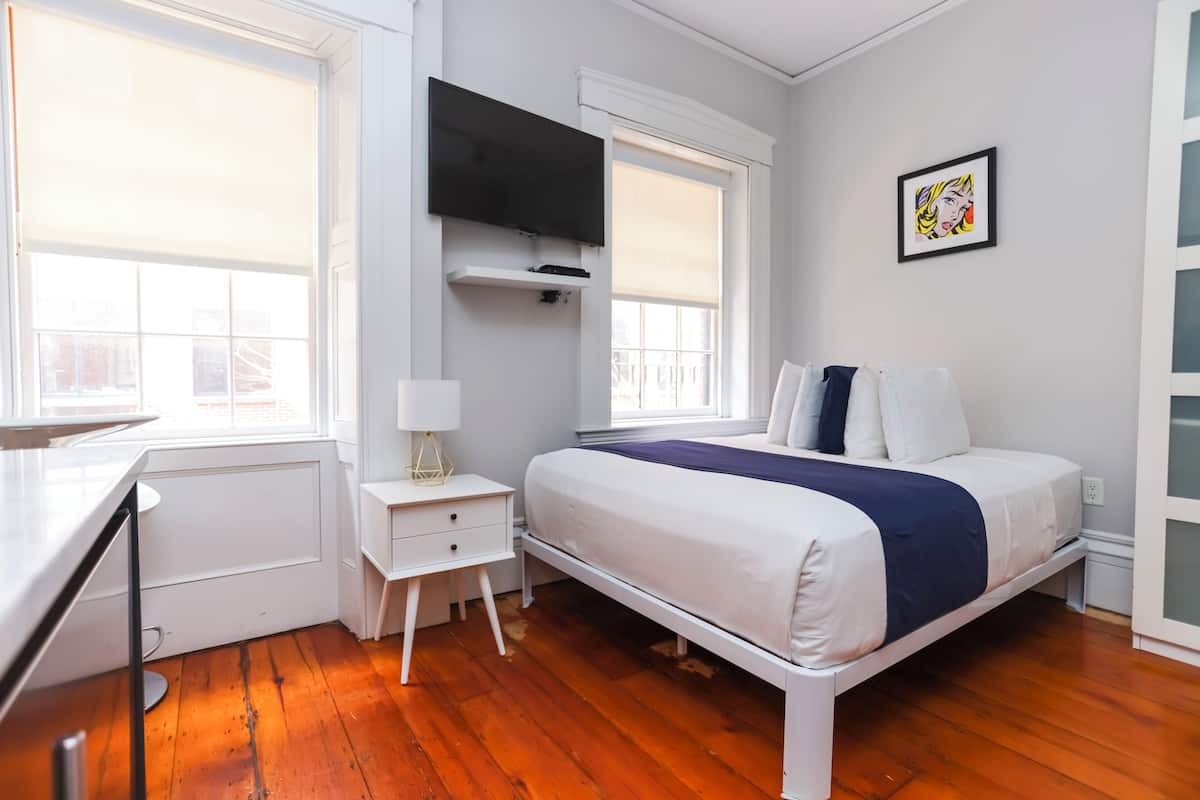 A bedroom with a bed, a desk, a TV, and a window.
