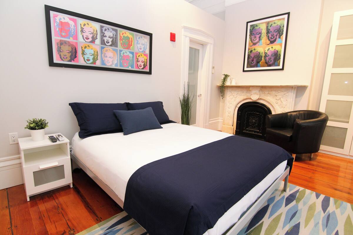 A neatly arranged bedroom with a bed, bedside tables, a fireplace, and artwork on the walls.