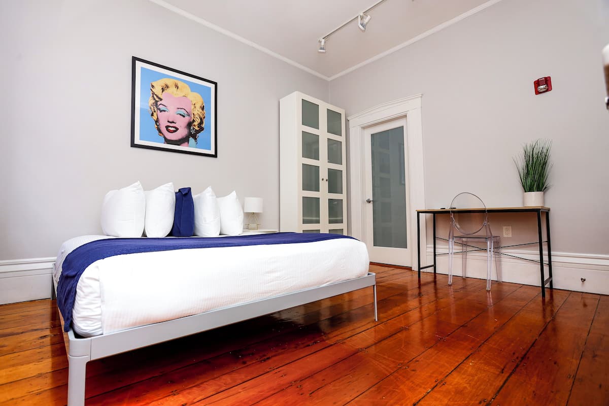 A bedroom with a bed, a white door, a small table with a chair, and a framed picture on the wall.