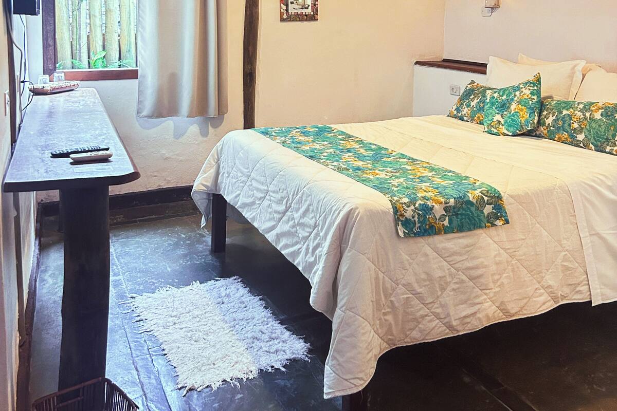 A bedroom with a bed, a desk, and a chair. There is a window with curtains, a wall-mounted light, and a small table with a remote control.