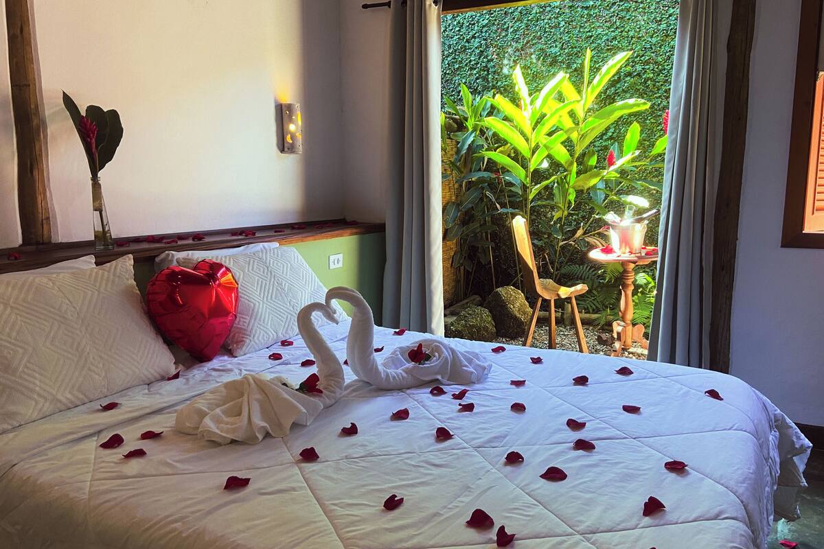 A neatly made bed with rose petals and a swan decoration, a red gift box, and a view of greenery through a window.