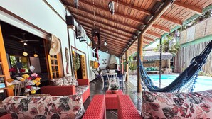 Un espacio de estar al aire libre techado con mobiliario de mimbre, una hamaca y una piscina al fondo.