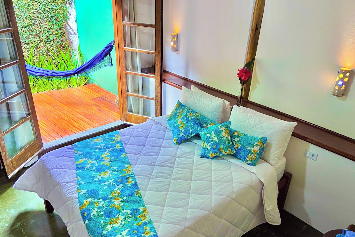 A bed with a floral bedspread and pillows, a wooden headboard, and a view of a wooden deck and green wall outside.