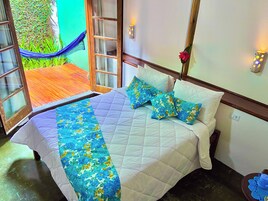 Een bed met een bloemenprint sprei en kussens, een houten hoofdbord, en uitzicht op een houten terras en een groene wand buiten.