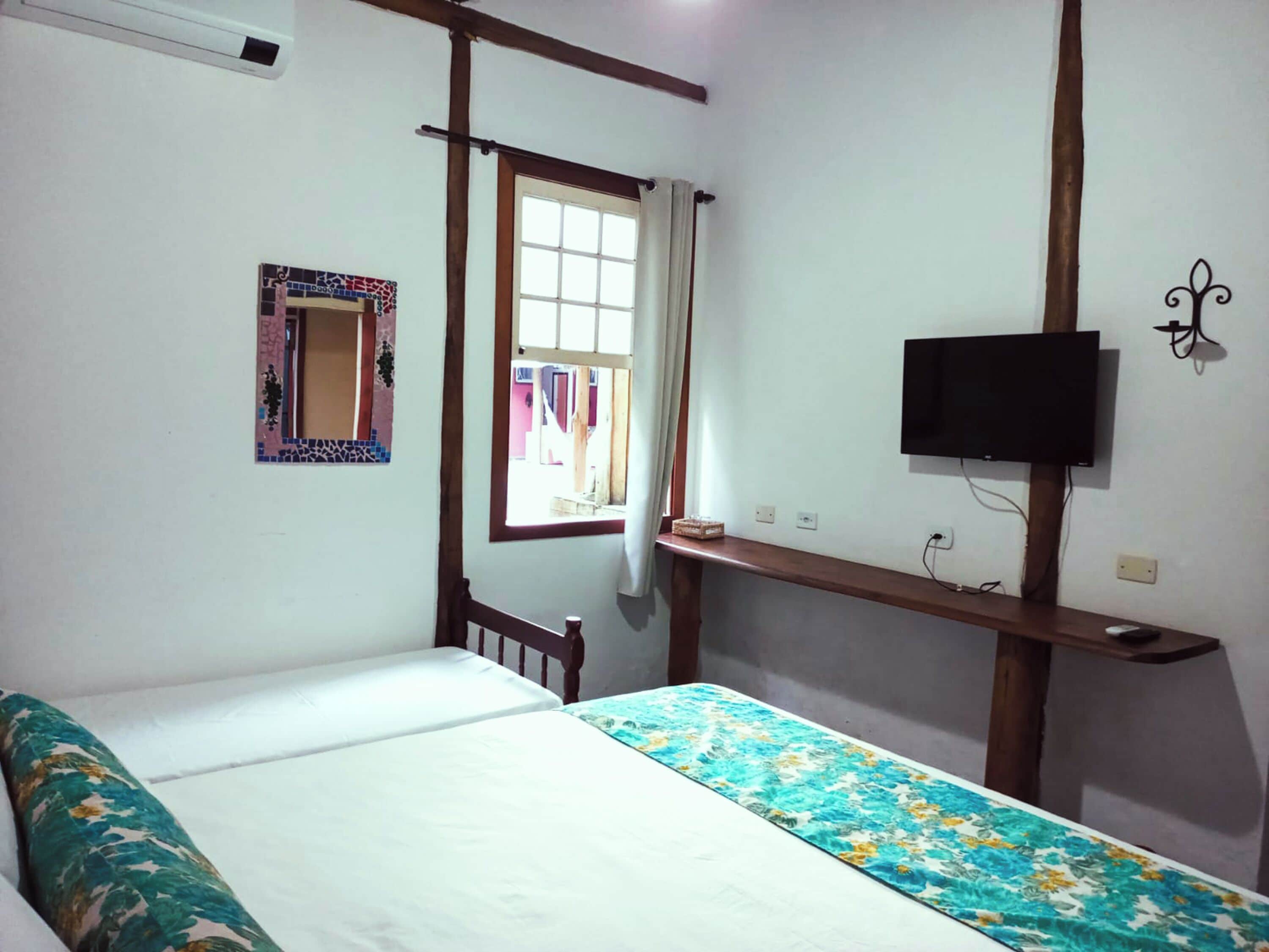 A bedroom with a bed, a TV mounted on the wall, a window with curtains, and a small shelf.