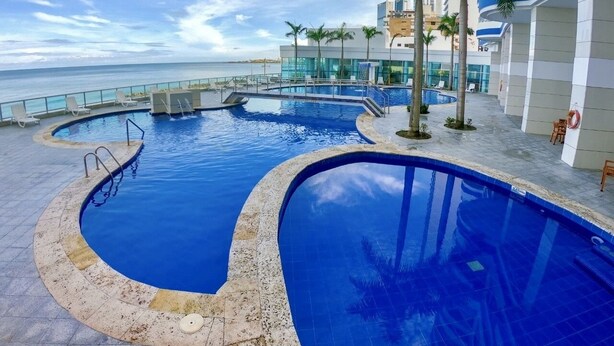 A swimming pool with a curved design, surrounded by palm trees and a building with a blue and white facade. - Enjoy the terrace and the best sunset and view of the city. (Cartagena de Indias)