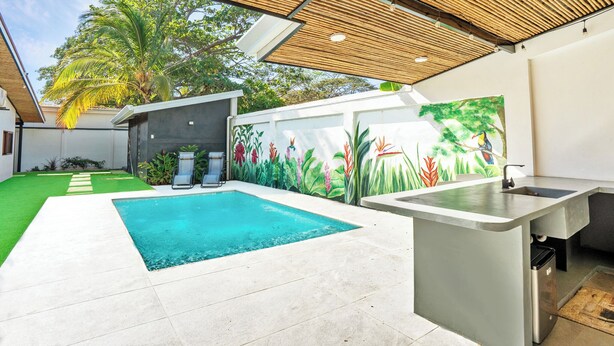 Un área moderna de cocina al aire libre con piscina, rodeada por un exuberante jardín y un mural decorativo.