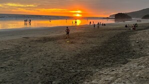 Escena de playa con gente caminando y jugando, un atardecer de fondo y un promontorio rocoso a la derecha.