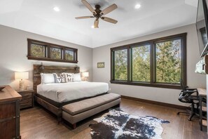 A bedroom with a bed, a desk, a chair, a ceiling fan, and large windows.