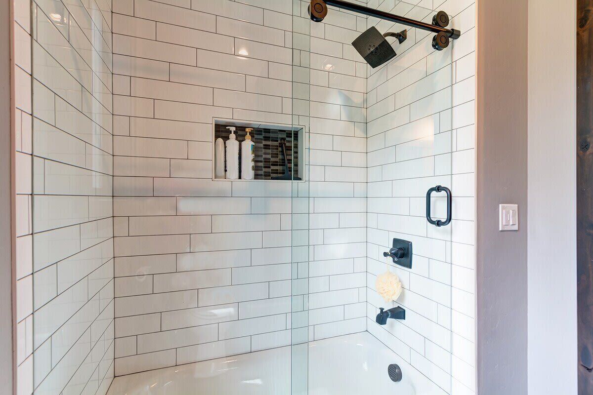 Un espace douche moderne avec des carreaux blancs, une pomme de douche protégée par une paroi de verre et une étagère intégrée pour les articles de toilette.