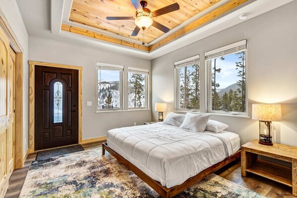 Un dormitorio con cama, ventilador de techo, ventanas amplias con vista a árboles y montañas, y una puerta de madera con un vitral.