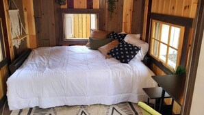 Un dormitorio rústico de cabaña de madera con una cama grande, dos ventanas y una planta colgante.