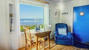 A room with a wooden table and chairs, a blue armchair, a view of the ocean, and a blue door. - Historic Oceanfront Cottage in Charming Nye Beach (Newport)