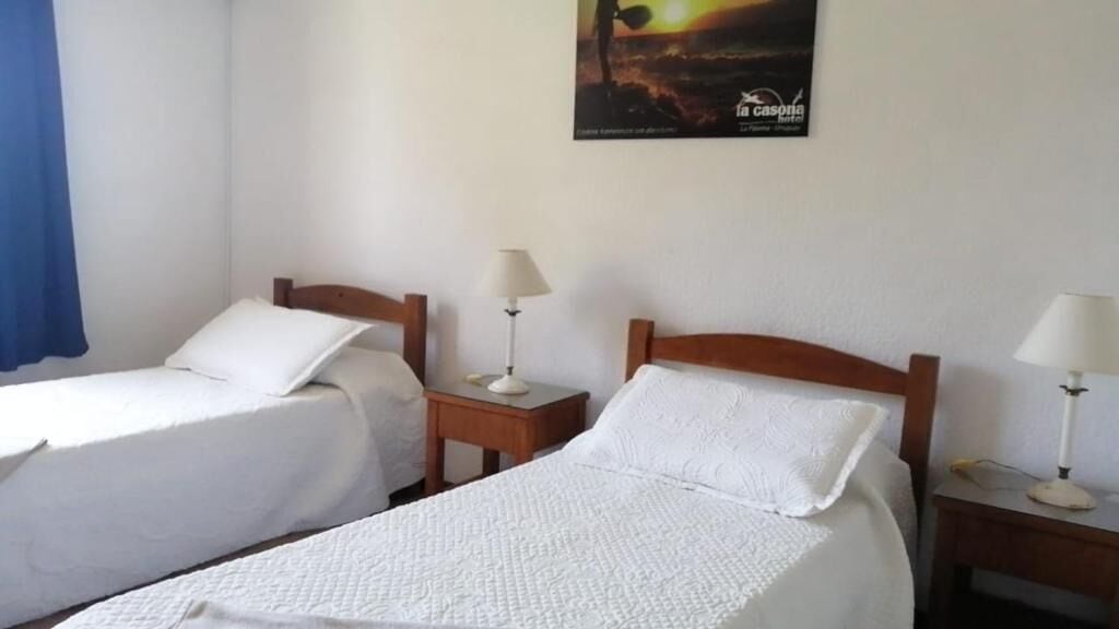 Une chambre d’hôtel avec deux lits simples, chacun doté d’une tête de lit en bois et de draps blancs. On y trouve des tables de chevet avec des lampes, ainsi qu’une affiche accrochée au mur au-dessus des lits.