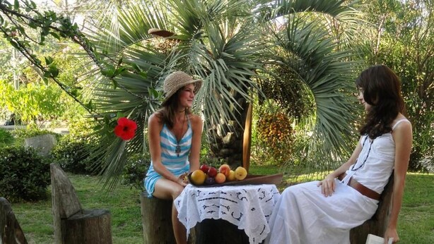 Due persone sedute su sgabelli di legno in un giardino, con un tavolo pieno di frutta di fronte.