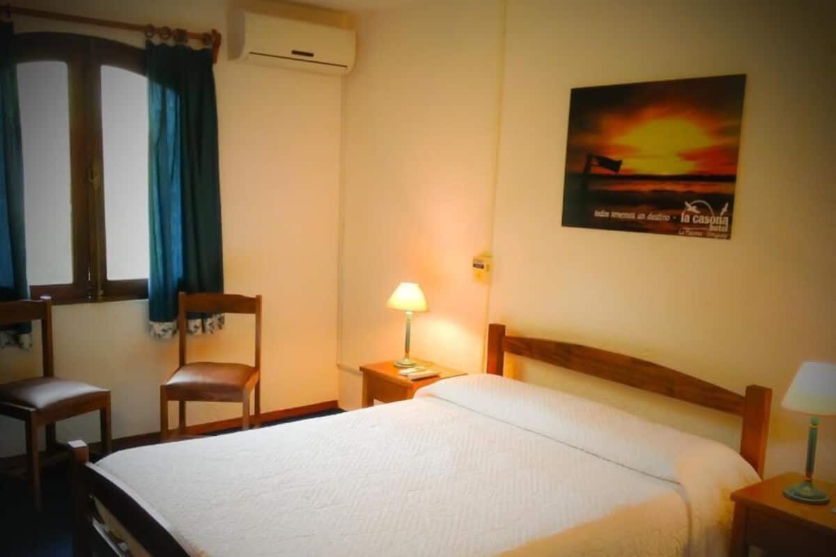 Una habitación de hotel con una cama, dos sillas, una mesita de noche con una lámpara, una ventana con cortinas y un cartel en la pared.