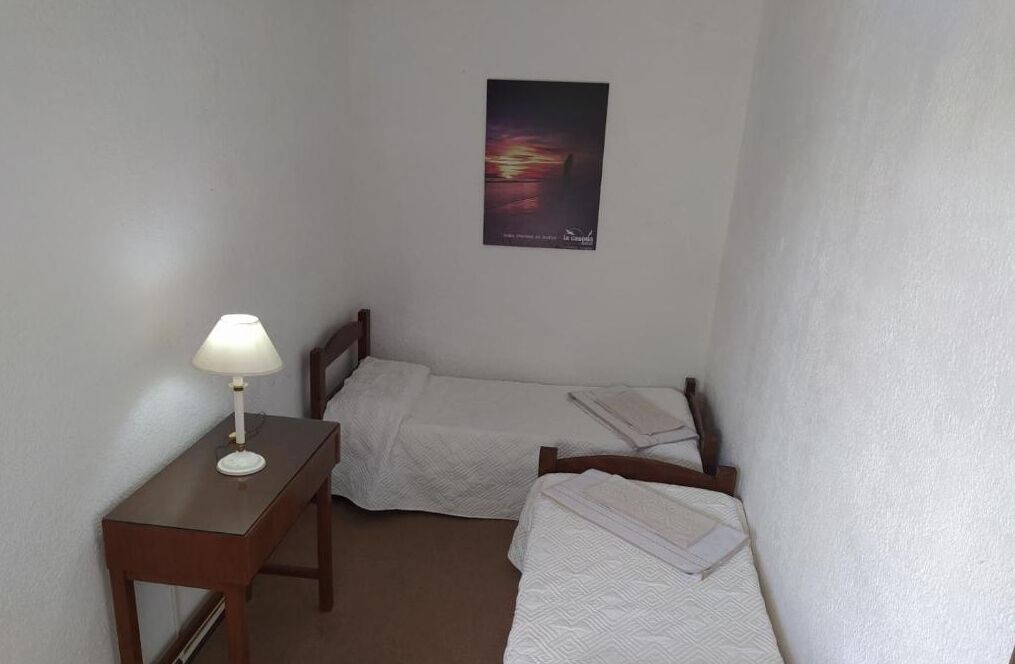 Um quarto pequeno e limpo, com duas camas individuais, uma escrivaninha de madeira com abajur e um quadro na parede.