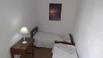 Um quarto pequeno e limpo, com duas camas individuais, uma escrivaninha de madeira com abajur e um quadro na parede.