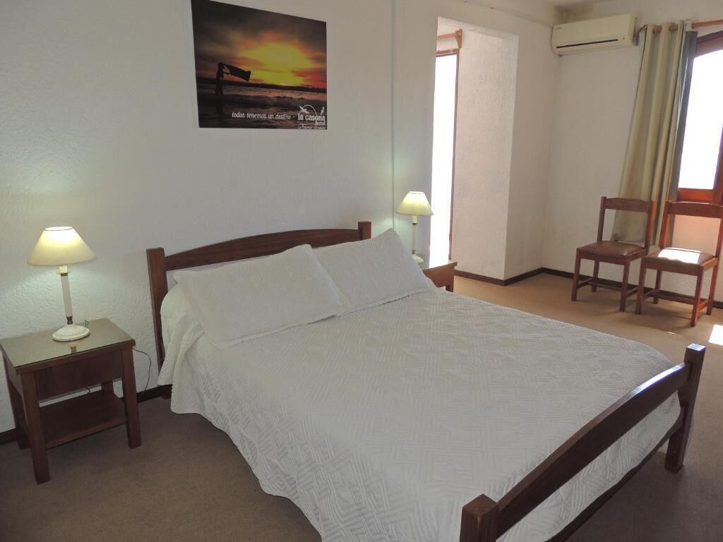 A neatly made bed with white linens, two bedside tables with lamps, a chair, and a wall-mounted air conditioner.