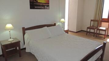 A neatly made bed with white linens, two bedside tables with lamps, a chair, and a wall-mounted air conditioner.
