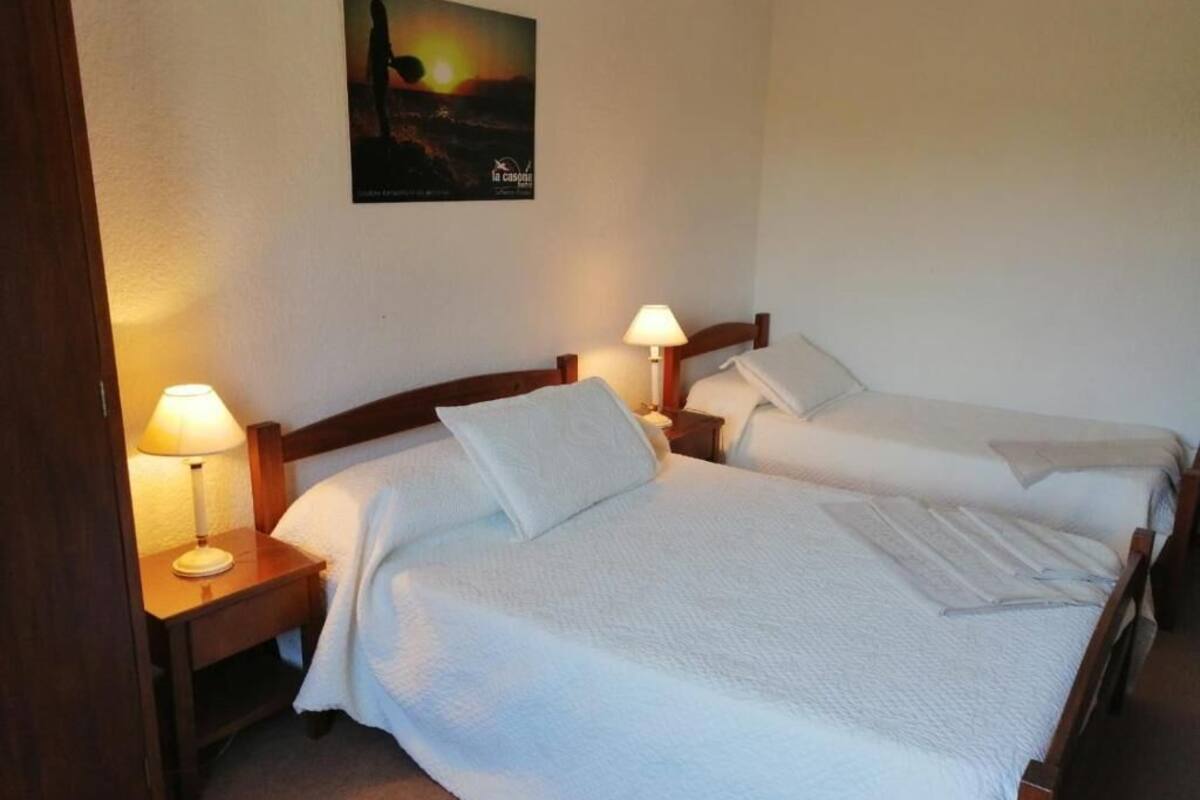 Habitación de hotel con dos camas individuales, cada una con cabecero de madera, mesita de noche y lámpara. Un cuadro enmarco cuelga de la pared sobre las camas.