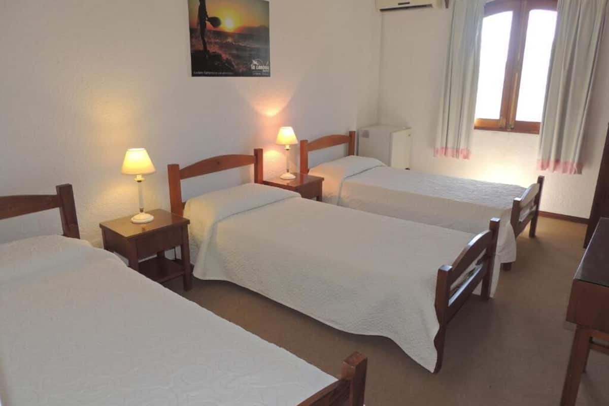 Habitación de hotel con tres camas individuales, cada una con cabecera de madera y mesita de noche con lámpara. Hay un cartel en la pared y un equipo de aire acondicionado sobre la ventana.