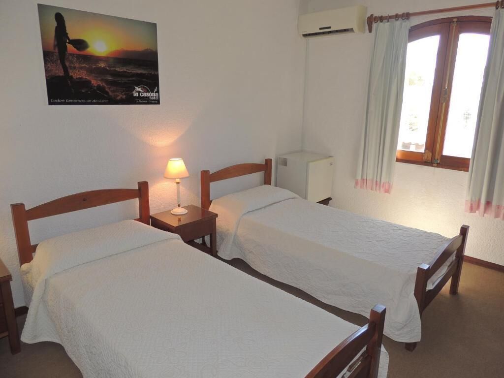 A room with two wooden beds, a bedside table with a lamp, an air conditioning unit, and a window with curtains.