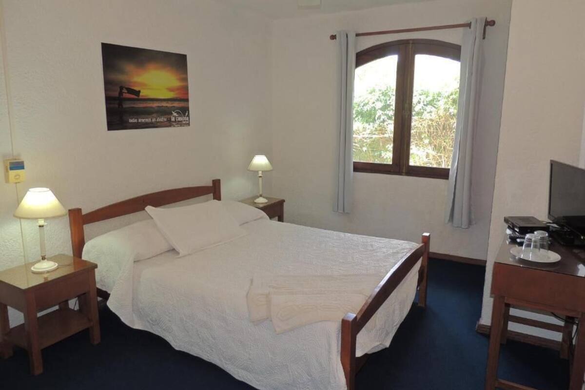 Un dormitorio con una cama, dos mesitas de noche con lámparas, un escritorio con silla, una ventana con cortinas y un cartel en la pared.