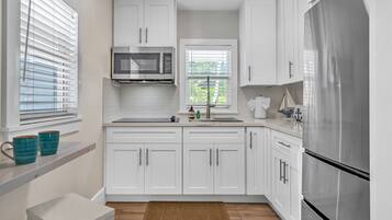 A modern kitchen with white cabinets, a stainless steel refrigerator, and a window with blinds.