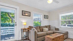 A living room with a sectional sofa, a wooden coffee table, and a side table with a lamp. There are framed pictures on the wall, a window with blinds, and a door leading outside. - Bungalow #4 - 2 Bed/1 Bath & Pool in Islamorada (Islamorada)