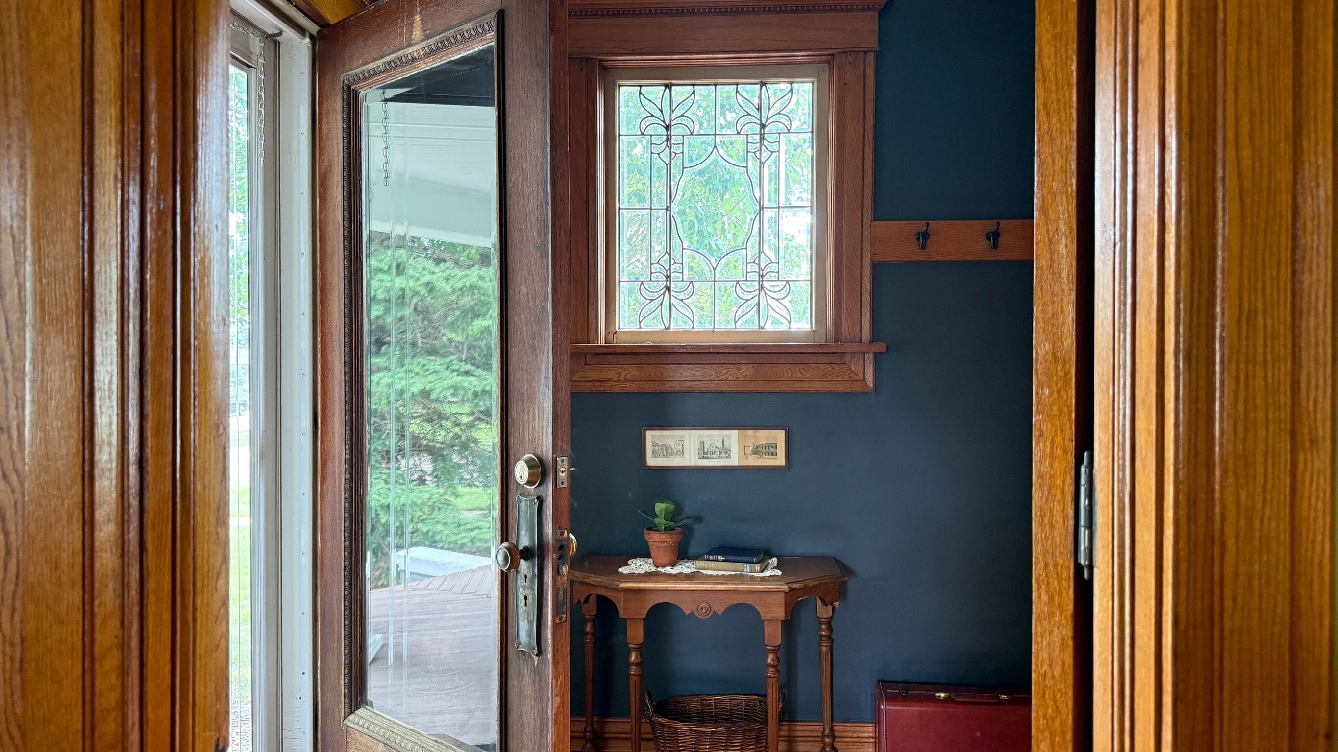 Une porte en bois avec un panneau de verre, une petite table sur laquelle est posée une plante, et une valise rouge.