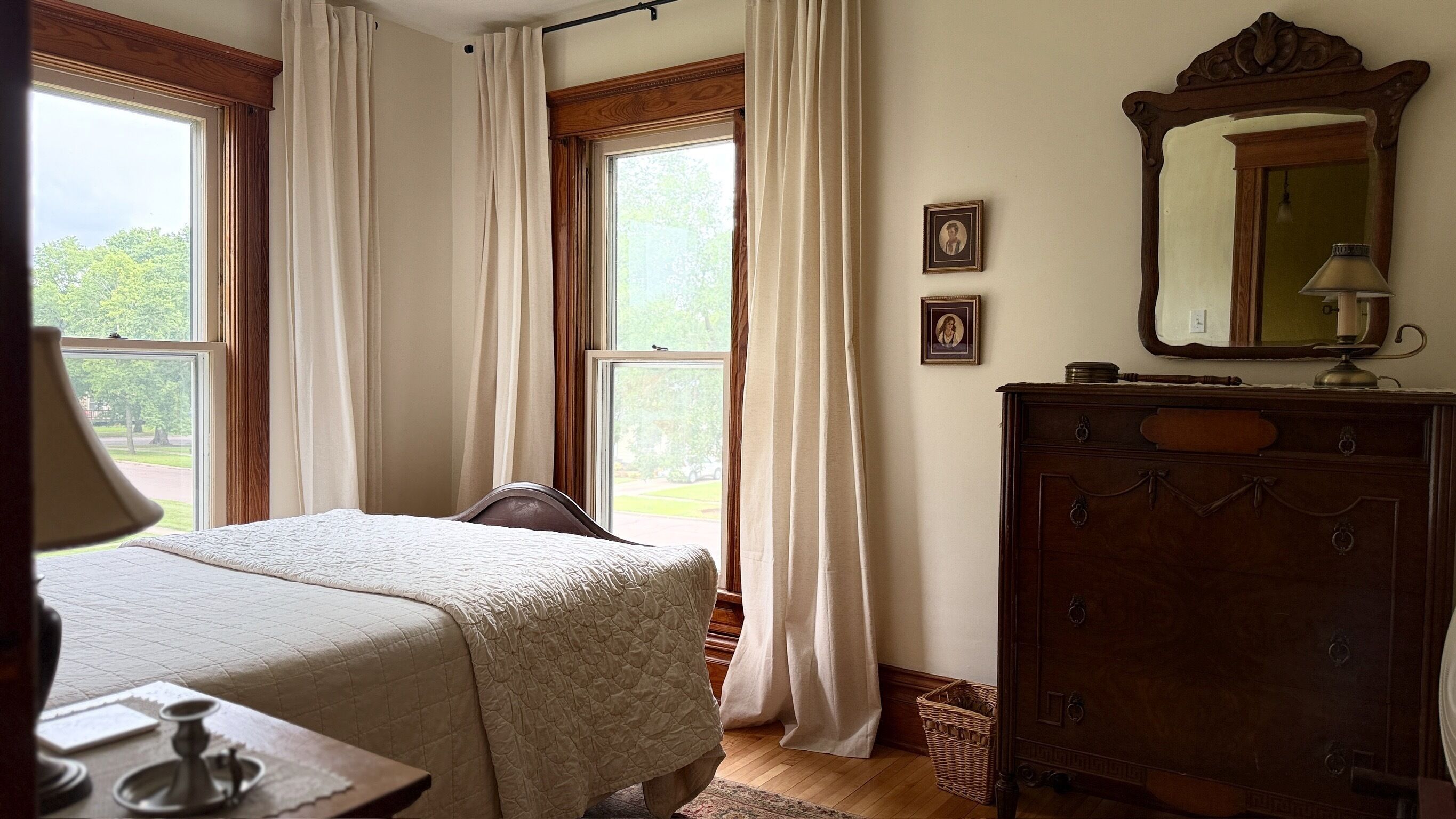 A bedroom with a bed, a dresser, a mirror, and two windows with curtains.