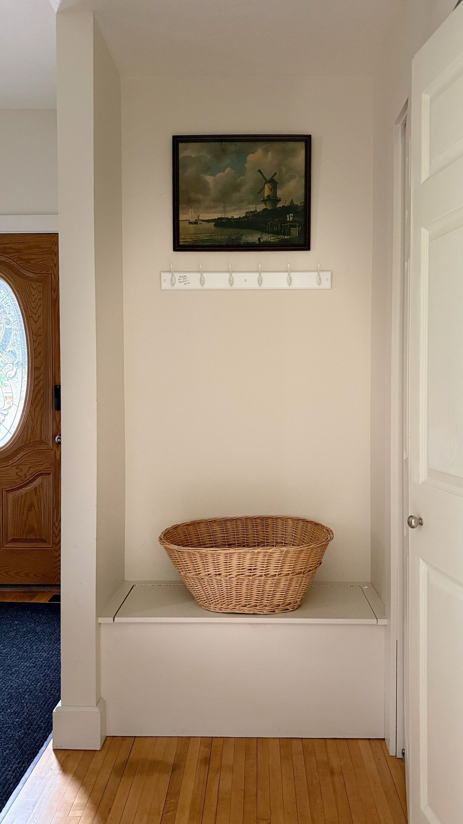 A room with a wooden door, a white wall, a framed painting, and a wicker basket on a shelf.