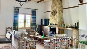 A spacious living room with exposed wooden beams, a stone fireplace, patterned sofas, and a dining table. - Rural house El Arado (Baena)