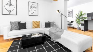 Een moderne woonkamer met een witte bank, een zwarte salontafel en een zwart vloerkleed. Twee ingelijste foto's hangen aan de muur, er staat een lamp en een plant.