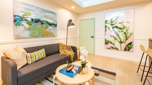 Una sala de estar moderna con un sofá gris, almohadones coloridos, una mesa redonda de centro con un libro y un cuenco decorativo, una lámpara de pie y dos cuadros grandes en las paredes.