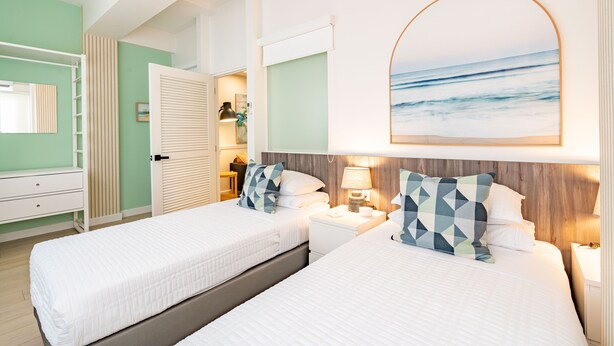 Una habitación de hotel con dos camas, un ventanal con vistas al océano, un cabecero de madera y un aparador blanco.