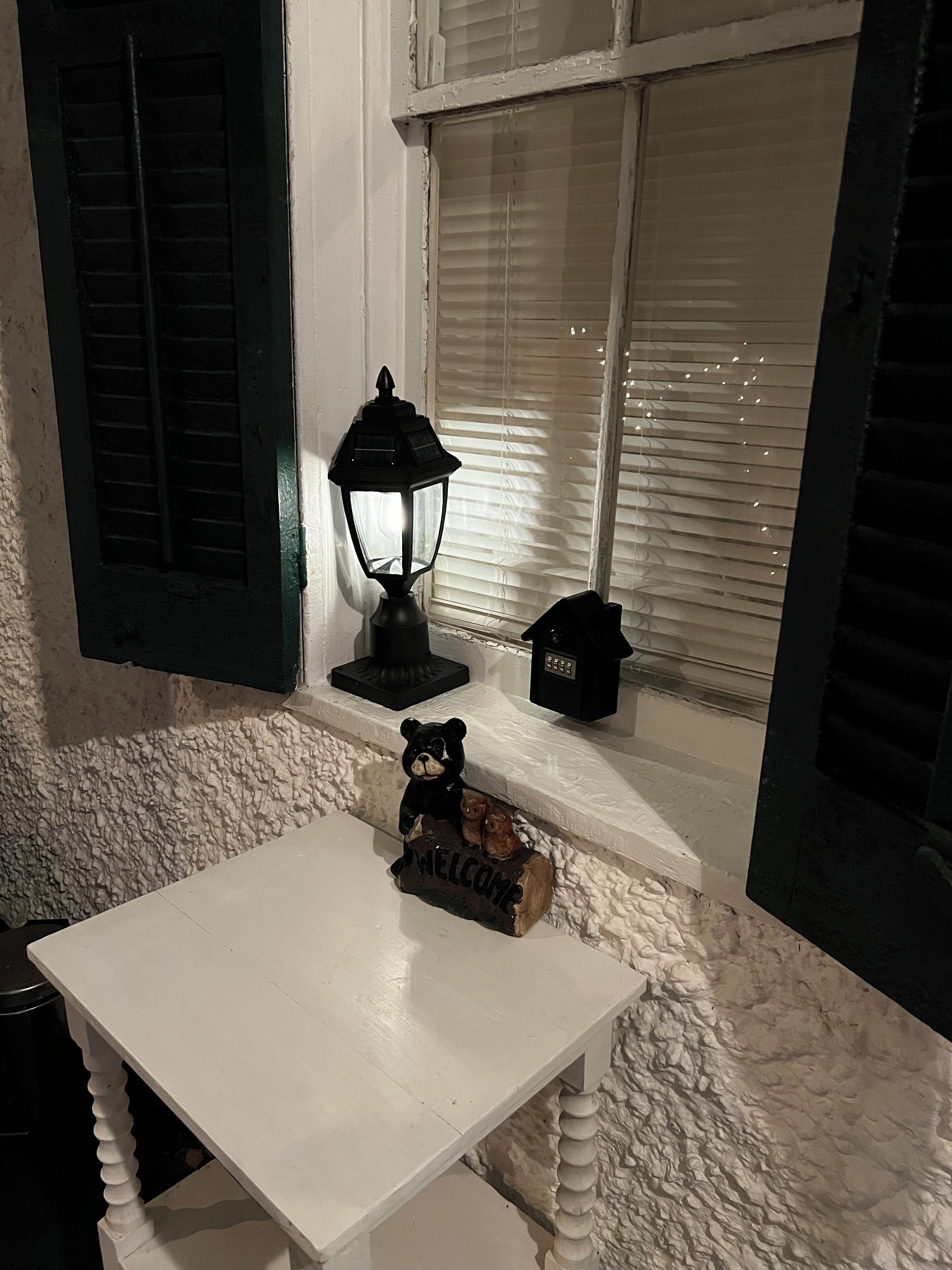 Ein weißer Tisch mit einer kleinen Bärenfigur, einem Willkommensschild, einer schwarzen Laterne und einem kleinen Hausmodell auf einem Fensterbrett.