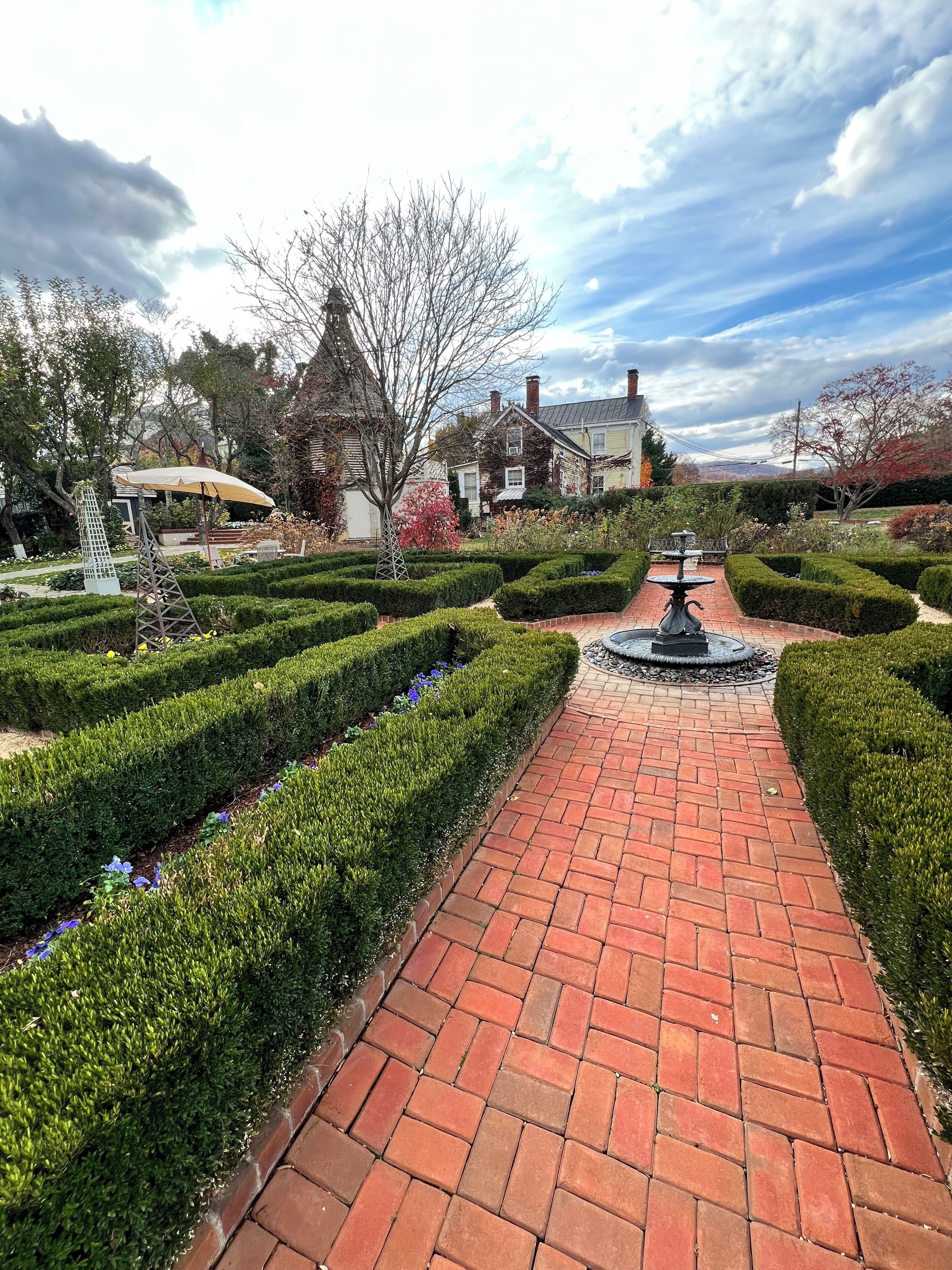 A brick pathway leading to a fountain in a well-maintained garden with trimmed hedges and a variety of plants.